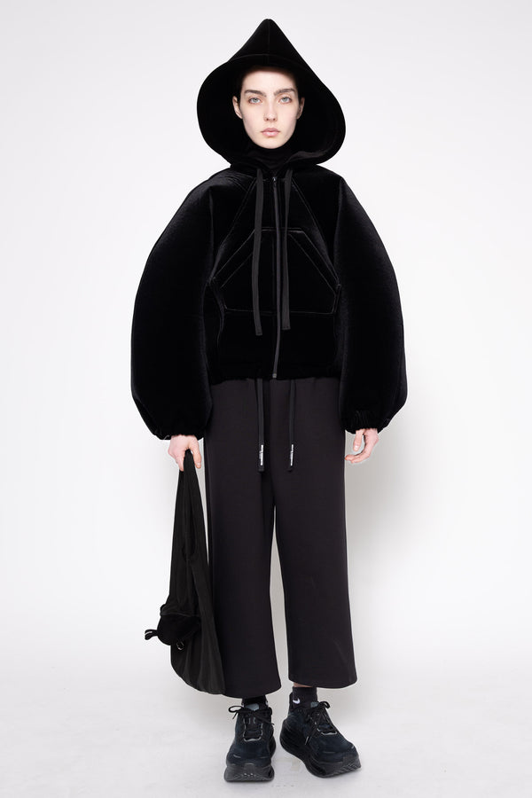 A model stands against a light gray studio background wearing a black hooded jacket with a fuzzy texture, black wide-leg pants, and black sneakers, while holding a black bag.