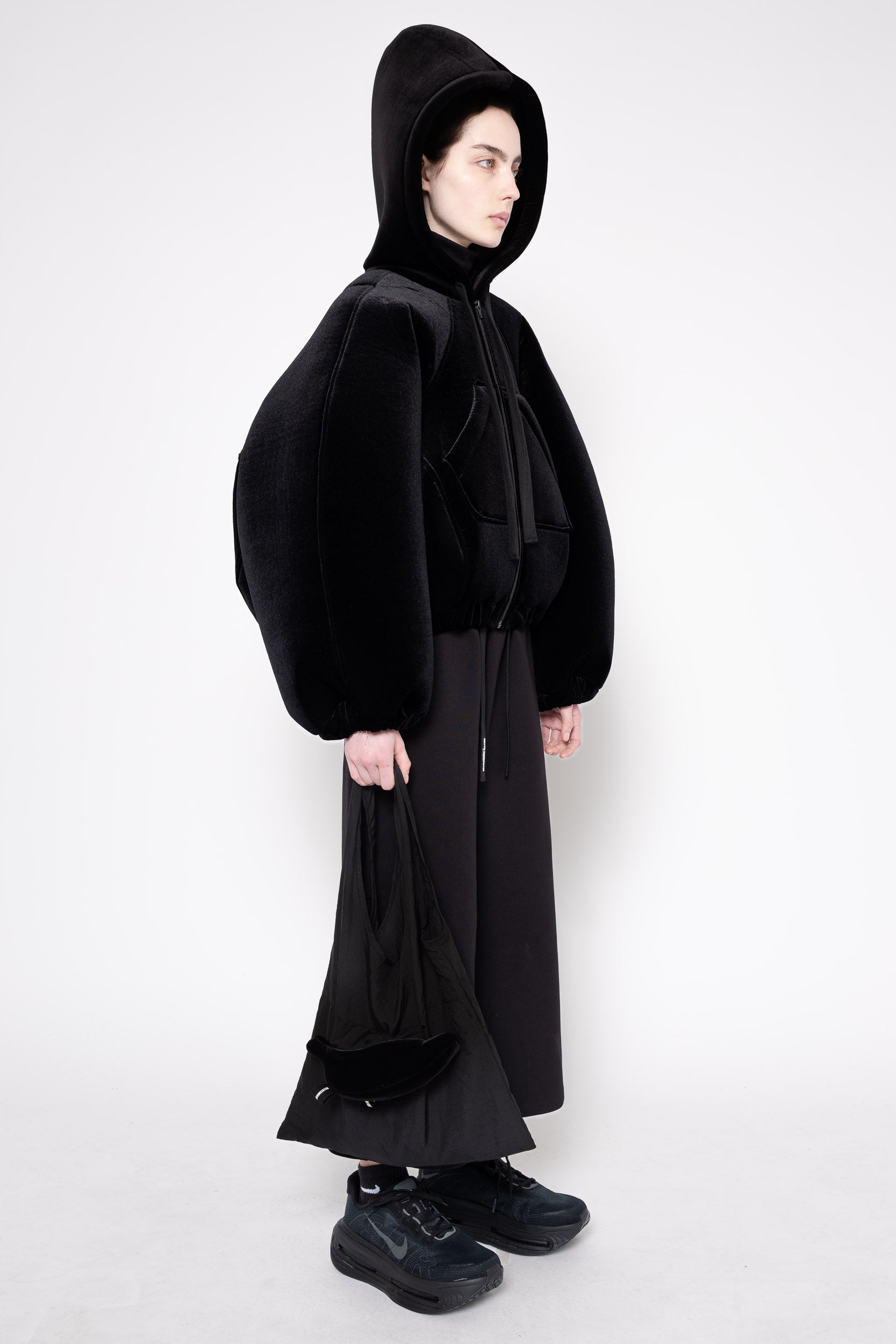 A model wearing a black hooded jacket with voluminous balloon sleeves, a black long skirt, and black sneakers, photographed against a plain light gray studio background.