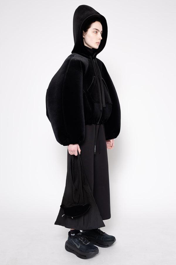 A model wearing a black hooded jacket with voluminous balloon sleeves, a black long skirt, and black sneakers, photographed against a plain light gray studio background.