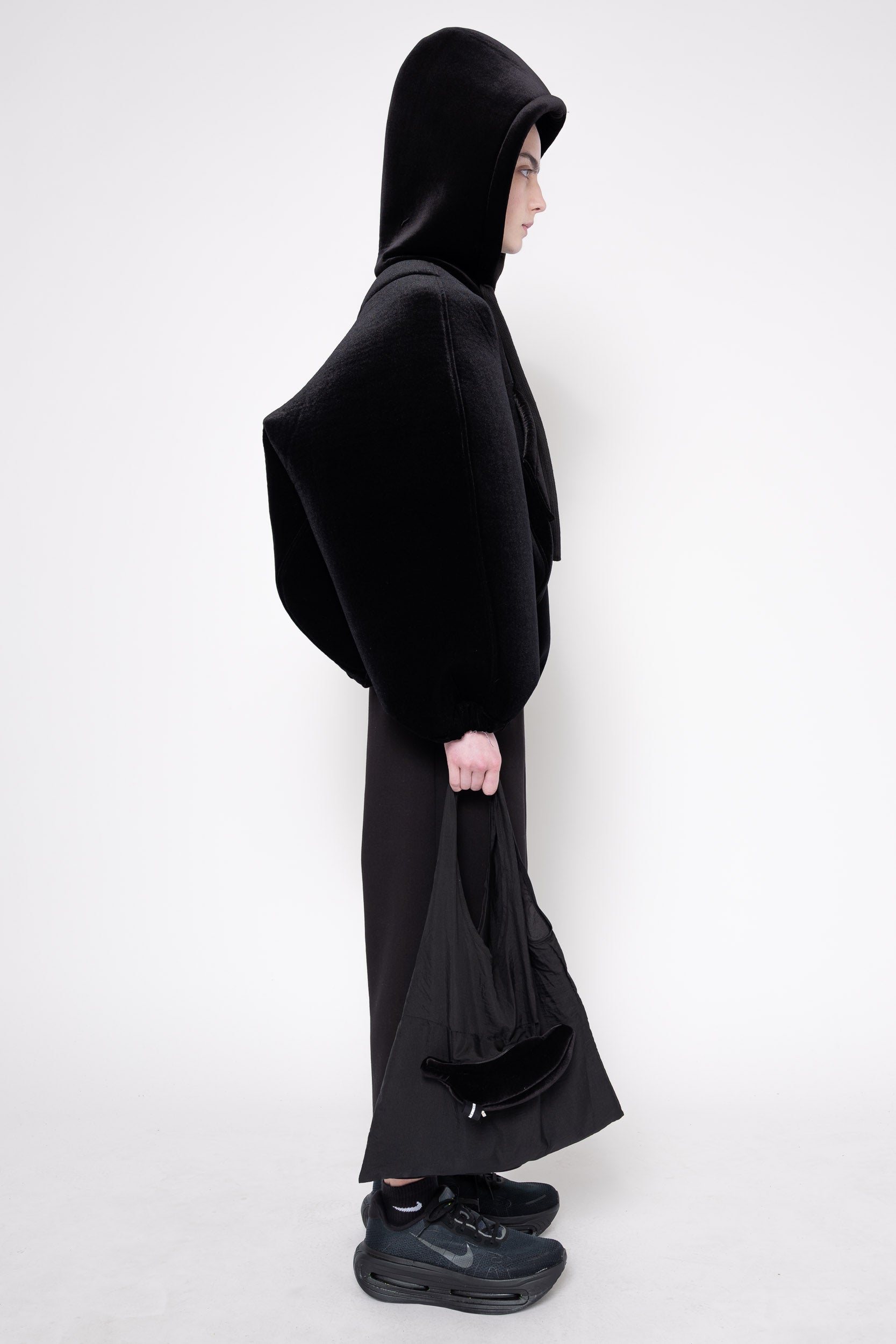 A model shown in profile wearing a black hoodie with wide draped sleeves and a long skirt-like drape, paired with black sneakers, against a plain light gray studio background.