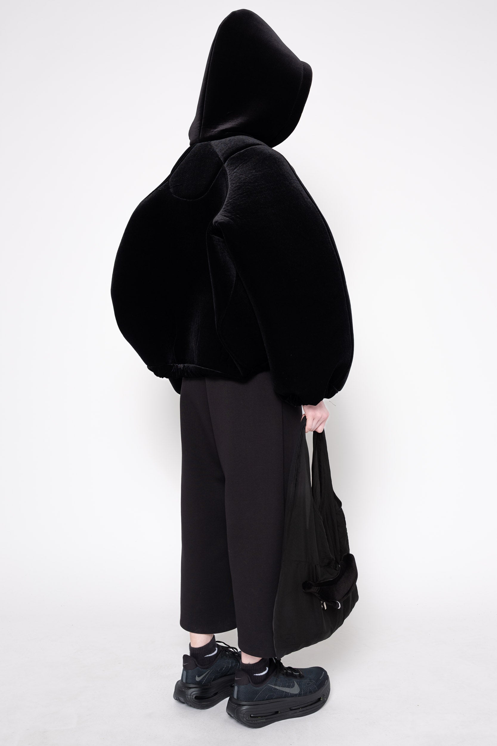 A model shown from the back wearing a black fuzzy cropped hoodie, black wide-leg trousers, black shoes, and holding a black tote bag against a plain white studio background.