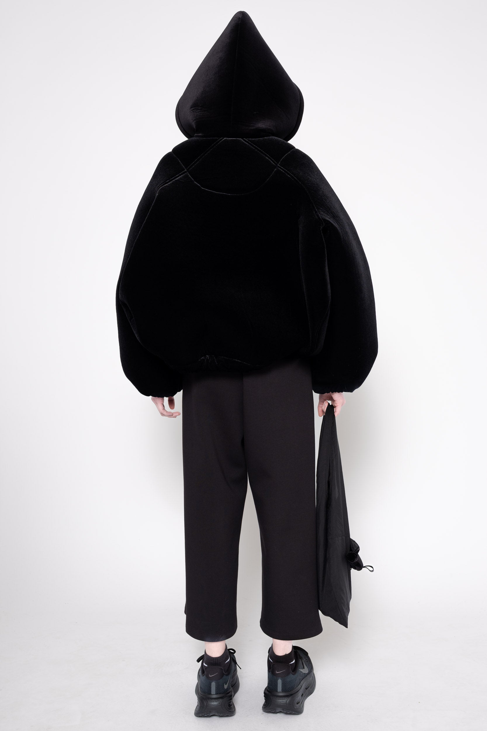A model shown from the back wearing a black hooded jacket made of plush velvet-like fabric, paired with black wide-leg cropped pants and black shoes, holding a black garment bag against a plain white studio background.
