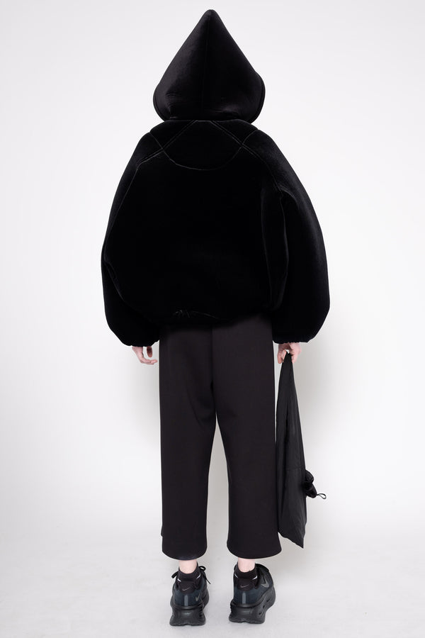 A model shown from the back wearing a black hooded jacket made of plush velvet-like fabric, paired with black wide-leg cropped pants and black shoes, holding a black garment bag against a plain white studio background.
