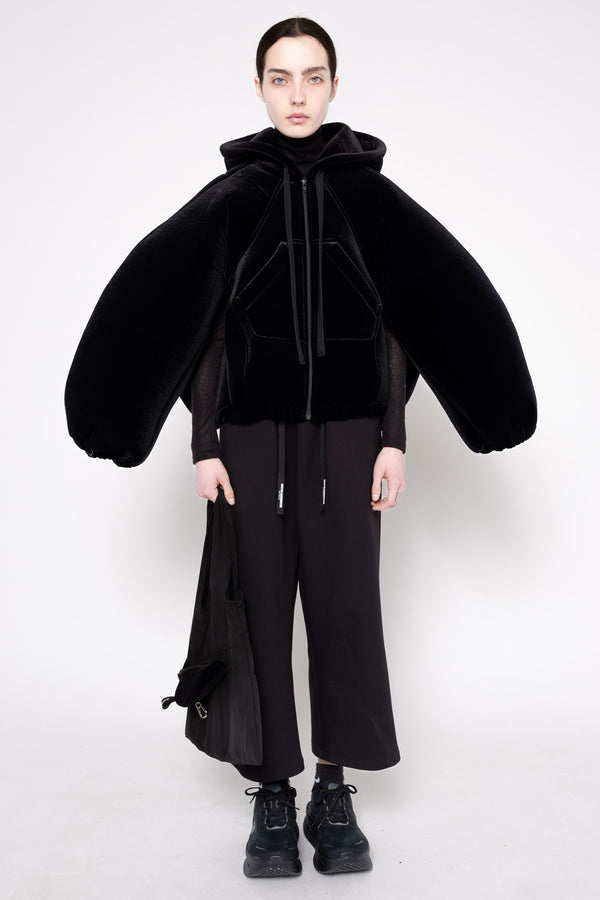 A model wearing a black futuristic hooded jacket with dramatic, oversized balloon sleeves, paired with black wide-leg trousers and black boots, shot against a plain white studio background.