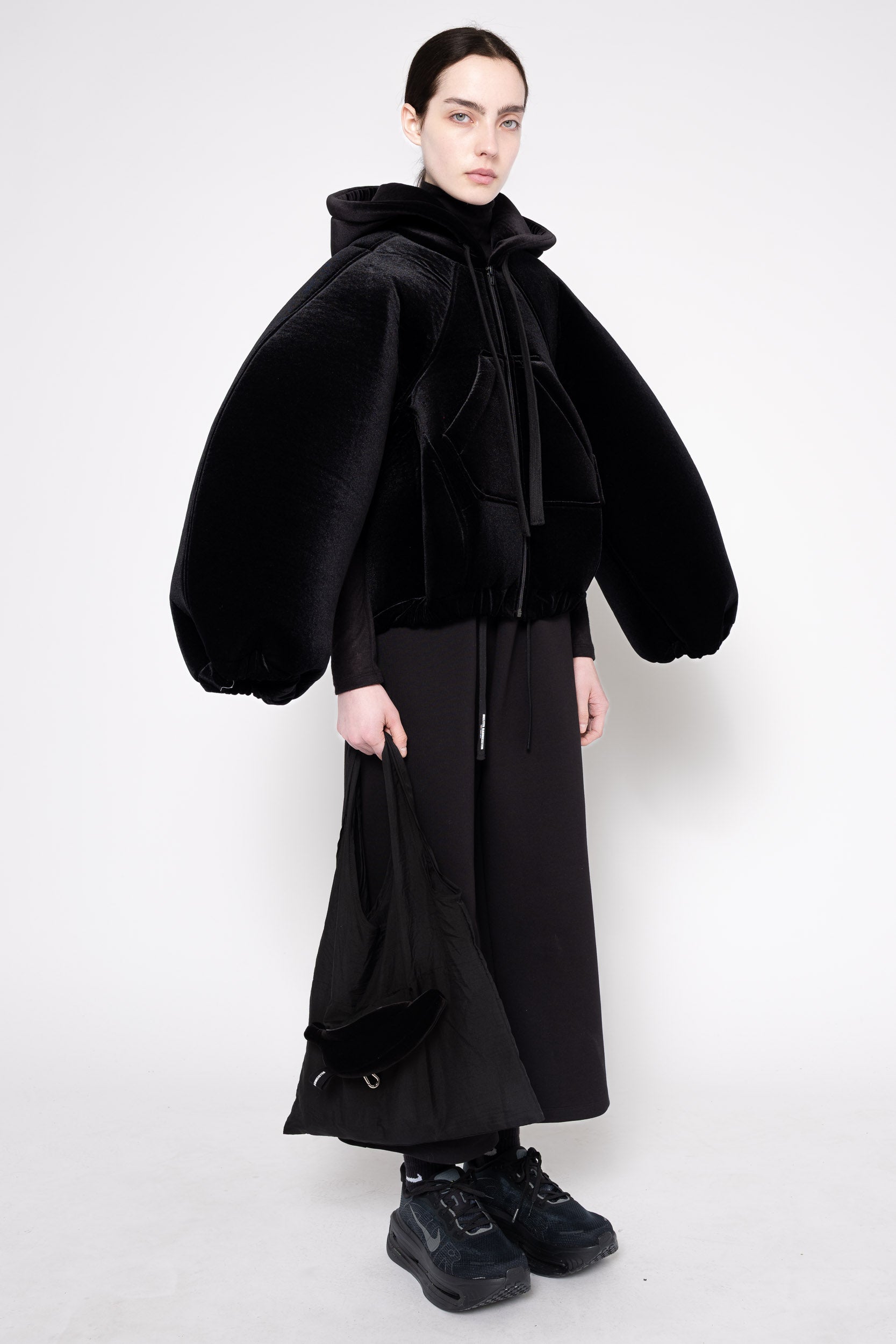 A model wearing a black hooded coat with oversized balloon sleeves, a black velvet tie detail at the neck, and a long black skirt, photographed against a plain light gray studio background.