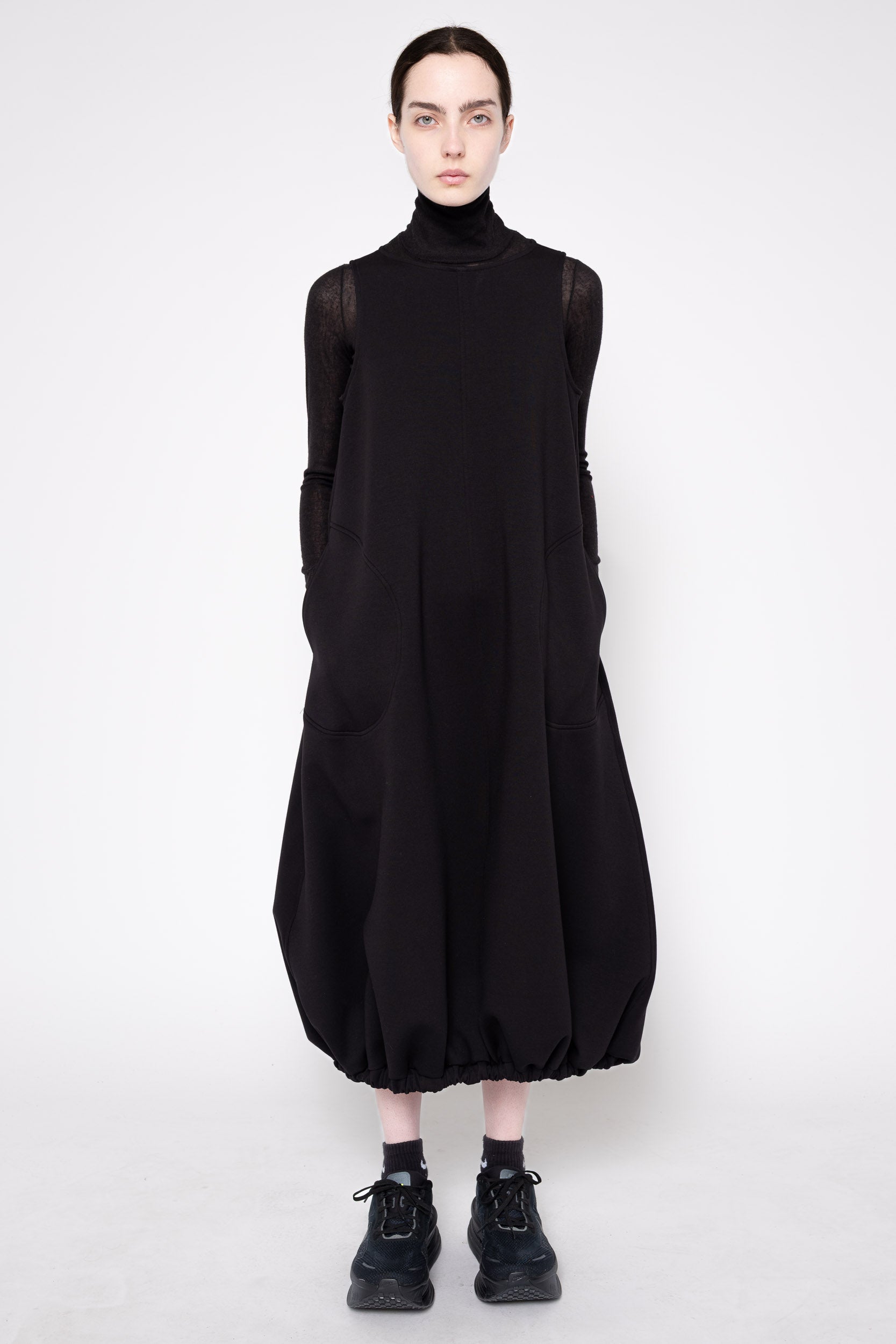 A model wearing a black long-sleeve turtleneck dress with voluminous balloon-style hem and cuffs, paired with black socks and sneakers, against a plain light gray studio background.