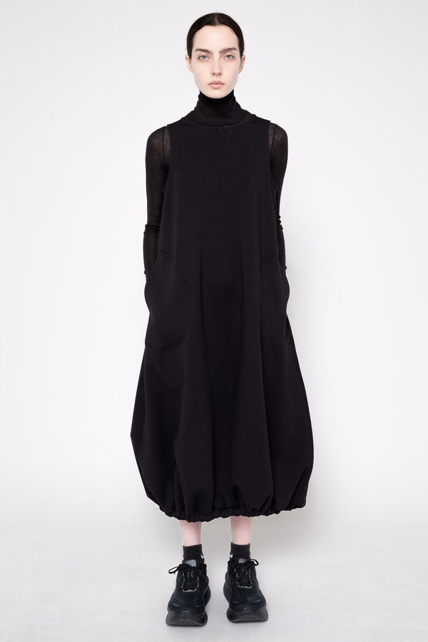 A model wearing a black long-sleeve turtleneck dress with voluminous balloon-style hem and cuffs, paired with black socks and sneakers, against a plain light gray studio background.