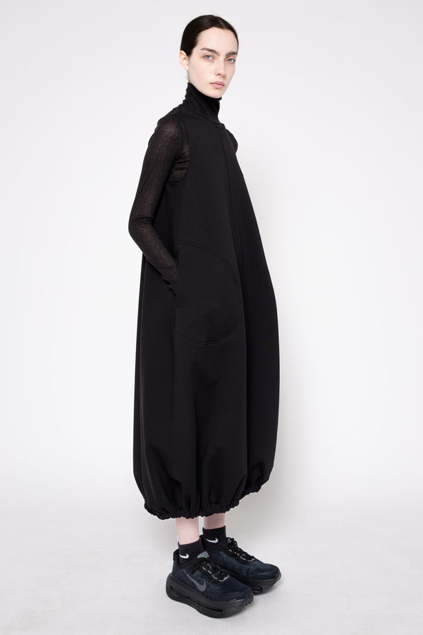 A model wearing a black long-sleeve turtleneck dress with voluminous gathered balloon hems and black sneakers, photographed against a plain light gray studio background.