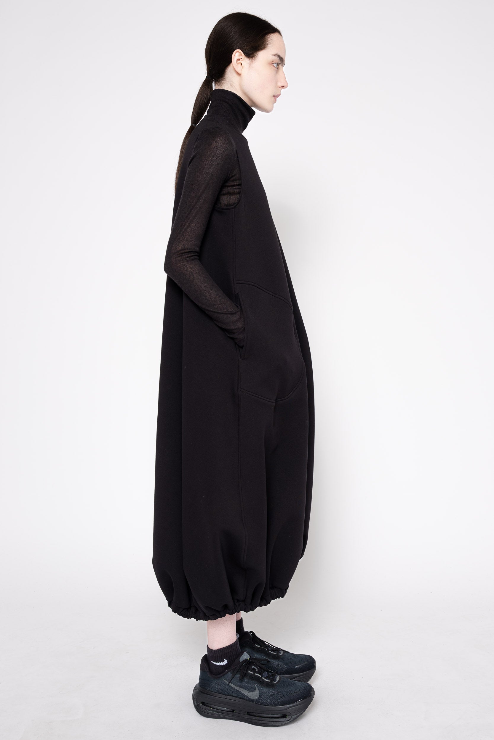 A model shown in profile wearing a long black turtleneck dress with sheer long sleeves and voluminous gathered legs, paired with black sneakers, against a plain light gray studio background.