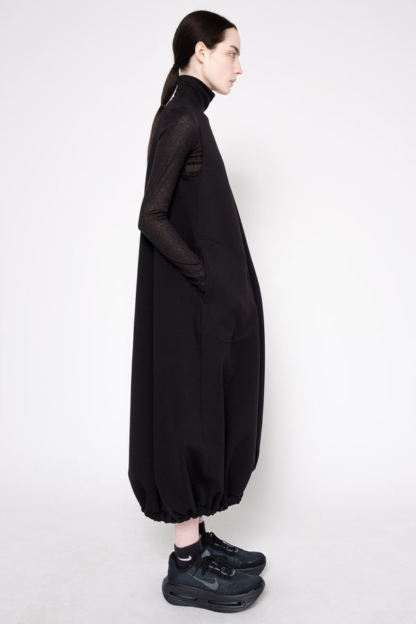 A model shown in profile wearing a long black turtleneck dress with sheer long sleeves and voluminous gathered legs, paired with black sneakers, against a plain light gray studio background.