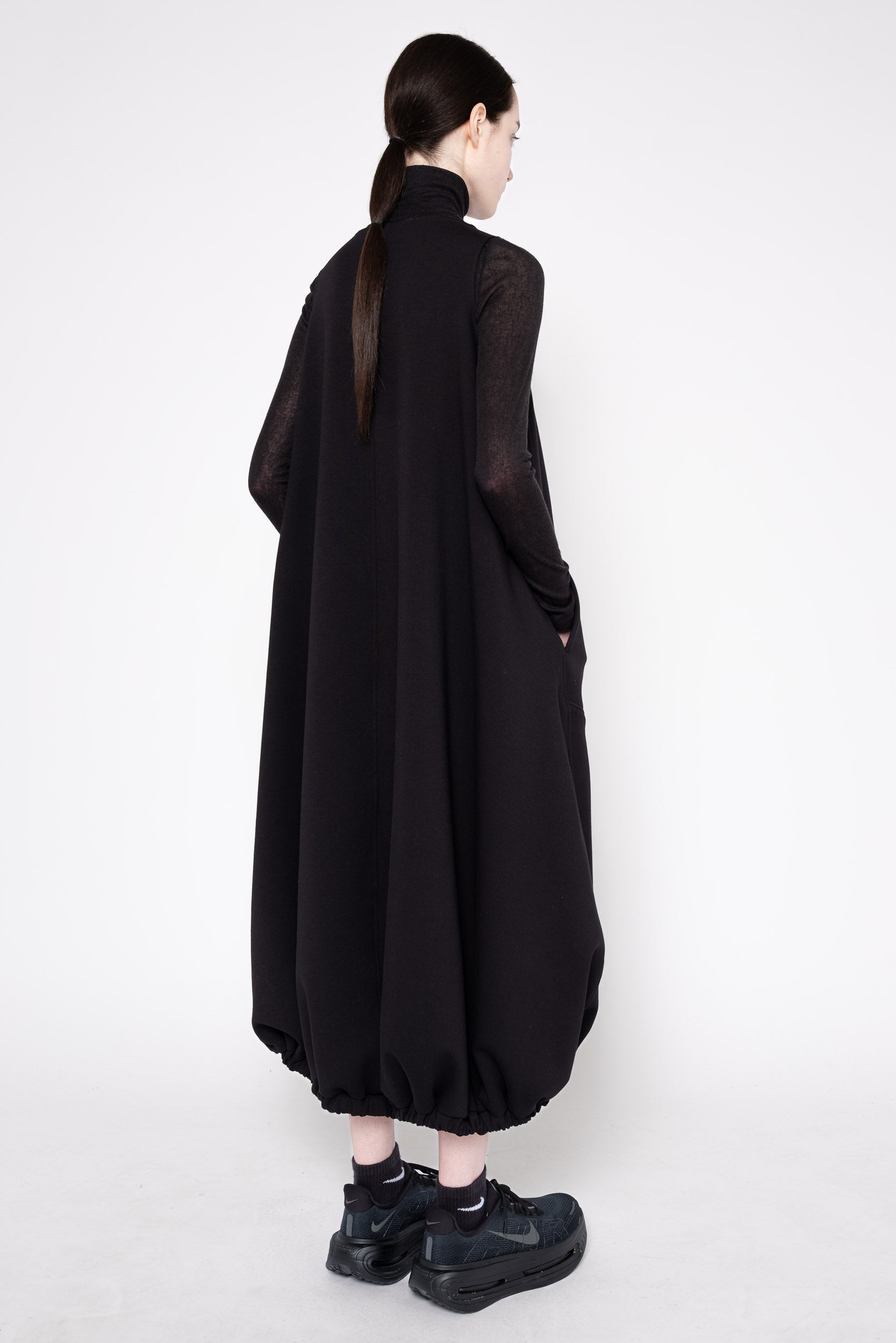 A model shown from the back wearing a long black high-neck dress with sheer long sleeves and gathered balloon hems, paired with black sneakers, against a plain white studio background.