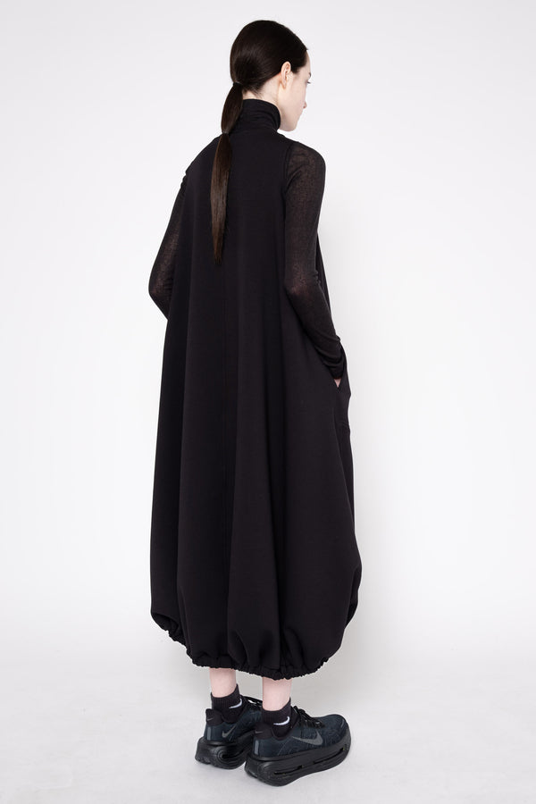 A model shown from the back wearing a long black high-neck dress with sheer long sleeves and gathered balloon hems, paired with black sneakers, against a plain white studio background.