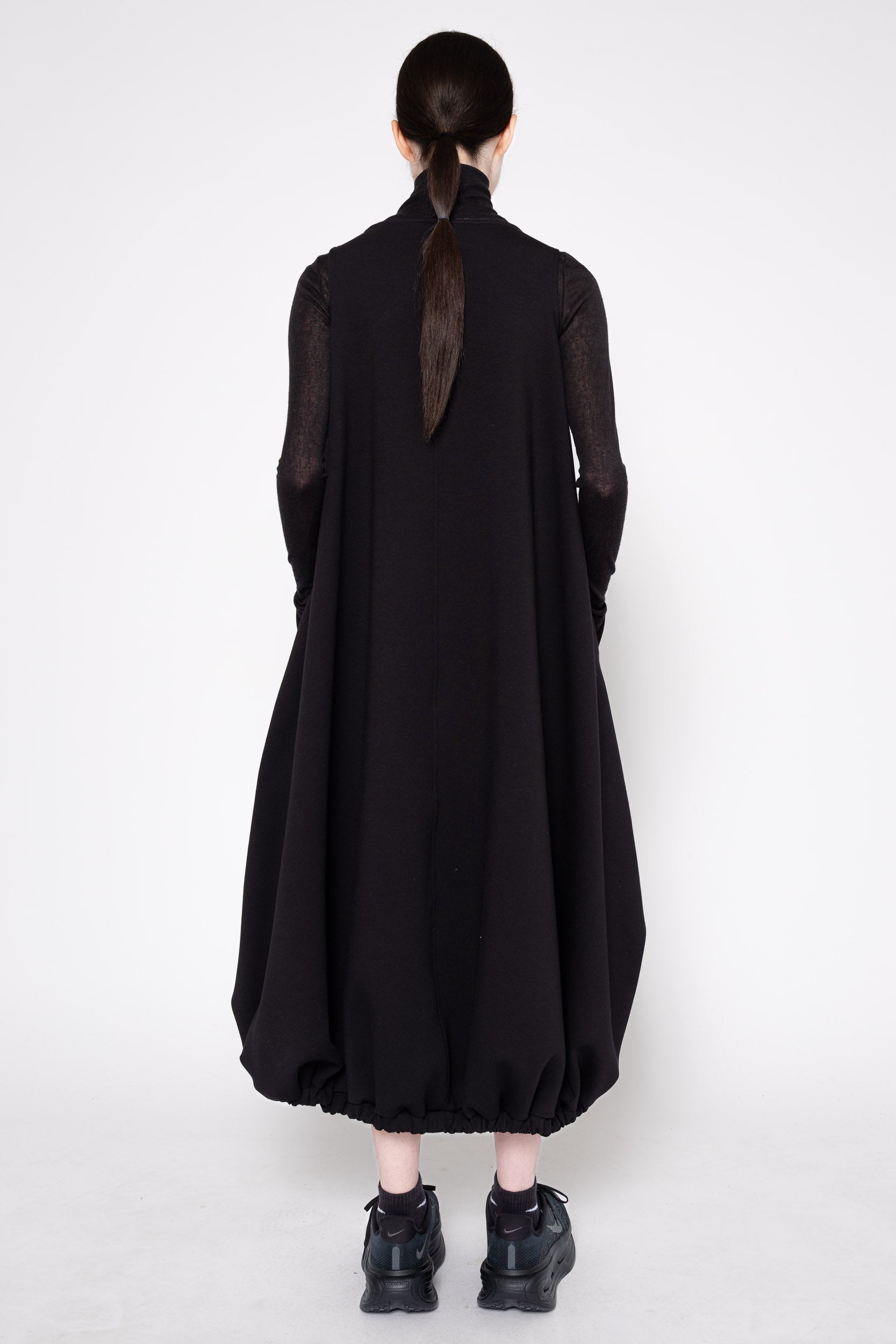 A model viewed from the back wearing a long black dress with sheer long sleeves and a high neck, paired with black shoes, against a plain light gray studio background.