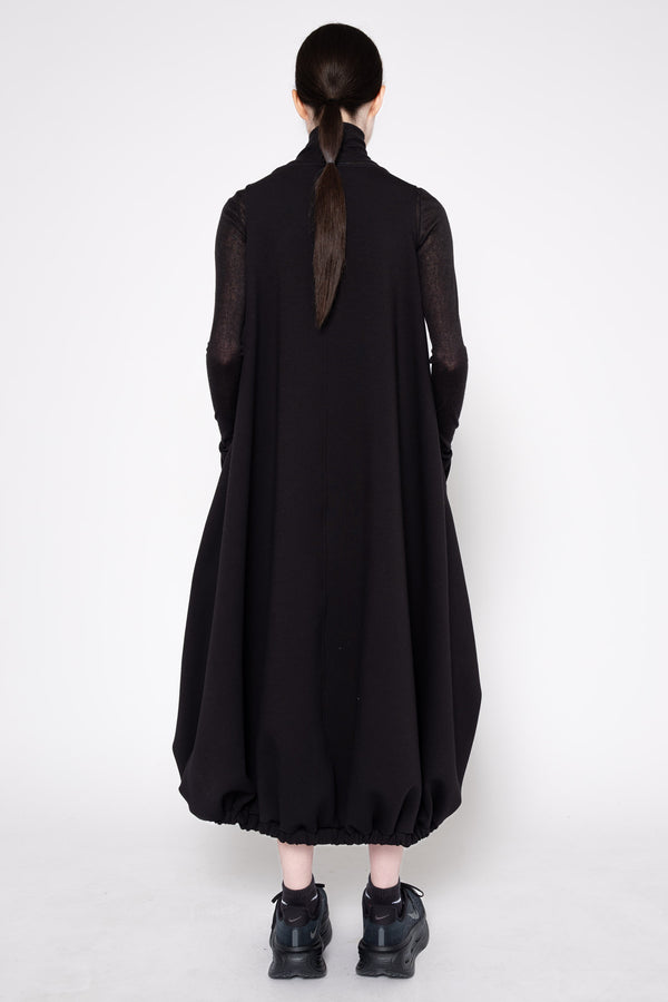 A model viewed from the back wearing a long black dress with sheer long sleeves and a high neck, paired with black shoes, against a plain light gray studio background.