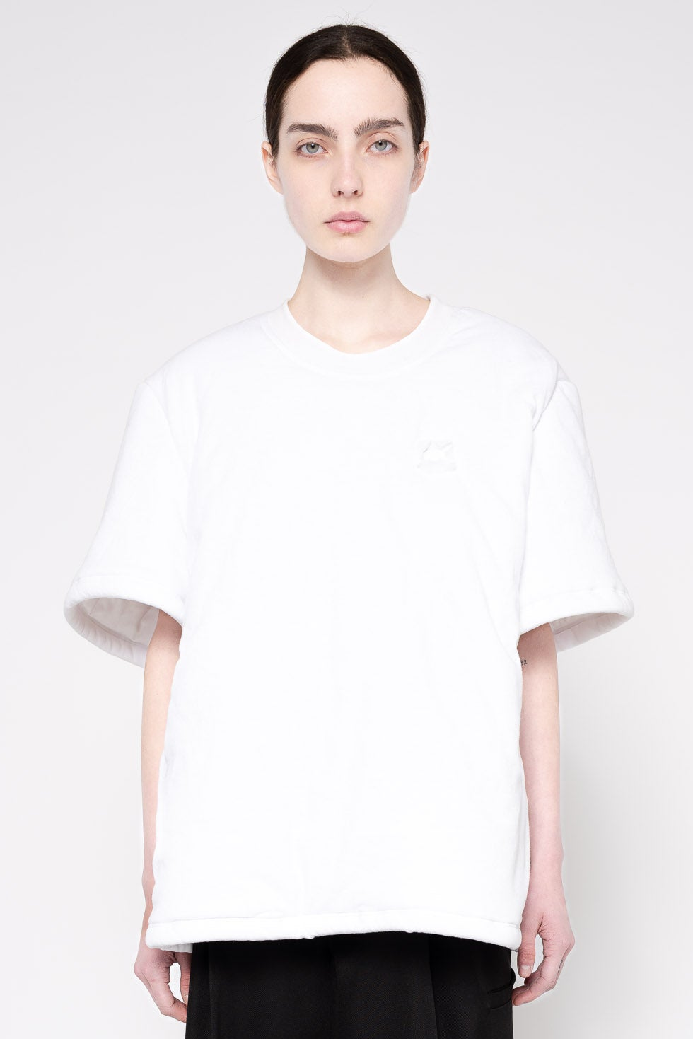 A model wearing an oversized white short-sleeve t-shirt with a subtle tonal embroidered logo on the left chest, paired with black pants, shot against a light gray studio background.