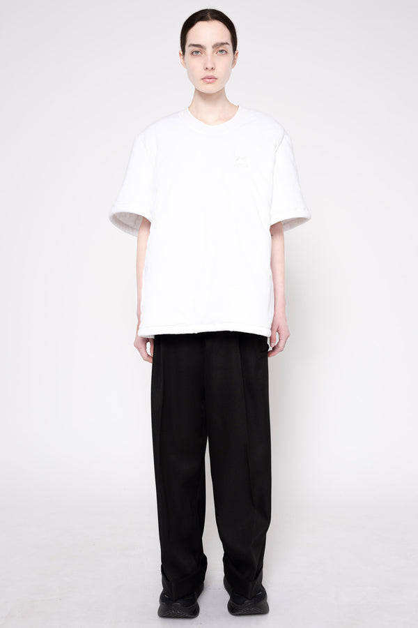A model stands against a light gray studio background wearing a loose white short-sleeve t-shirt and loose black wide-leg trousers.