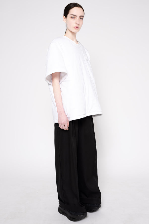 A model wearing an oversized white short-sleeve t-shirt and loose black wide-leg trousers, standing against a plain light gray studio background.