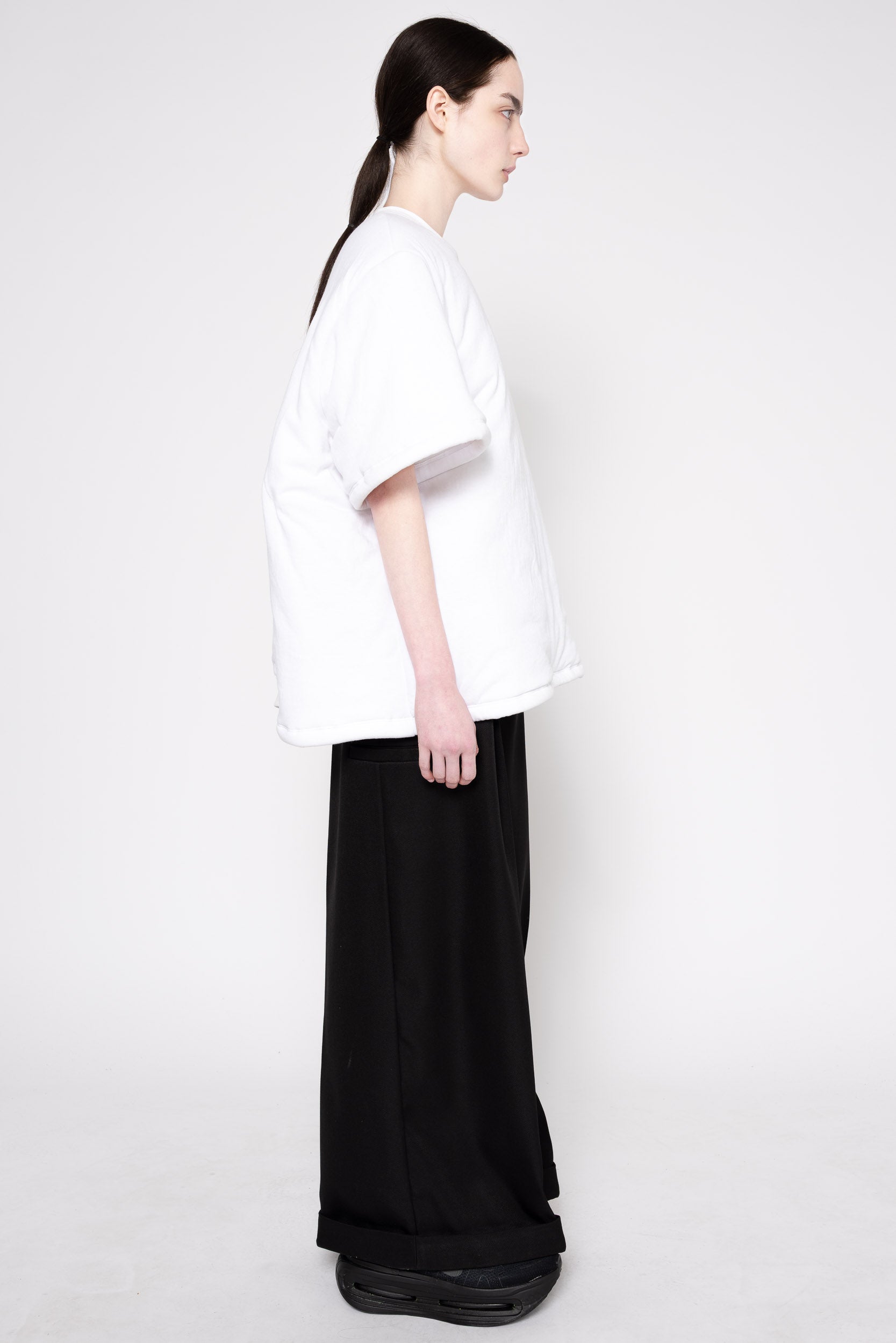 A model shown from the side profile wearing an oversized white short-sleeve t-shirt and wide-leg black trousers, standing against a plain light gray studio background.