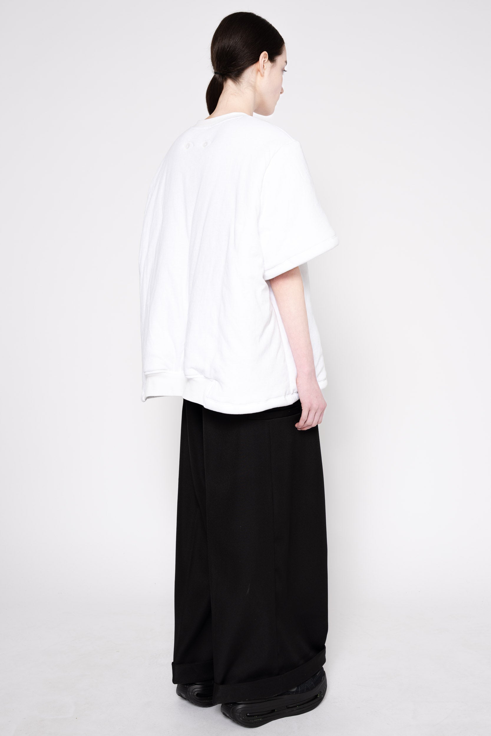 A model shown from the back wearing an oversized white short-sleeve top and wide-leg black trousers against a plain light gray studio background.