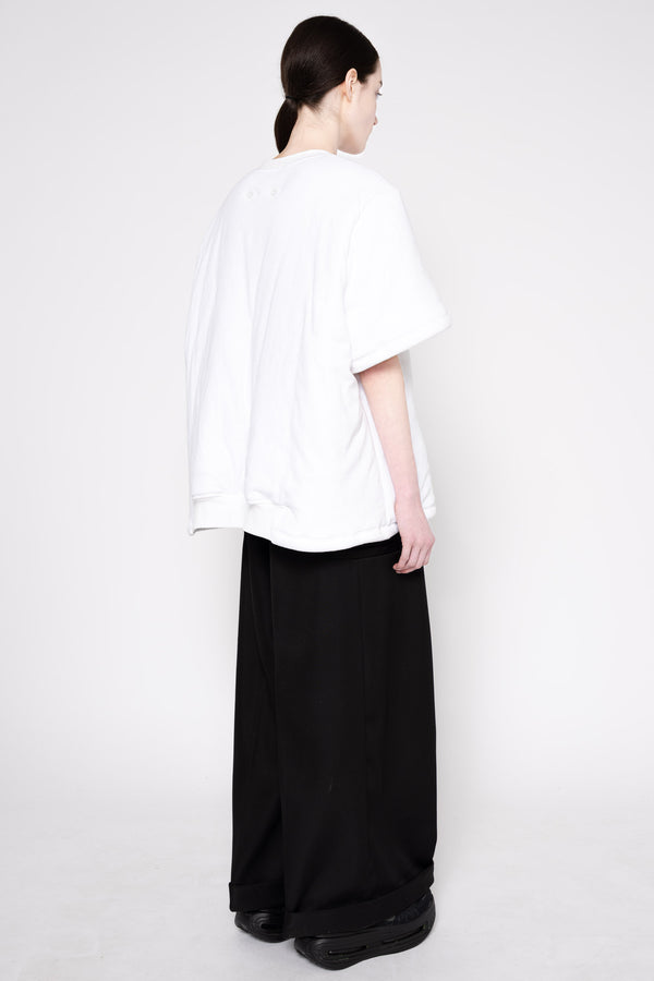 A model shown from the back wearing an oversized white short-sleeve top and wide-leg black trousers against a plain light gray studio background.