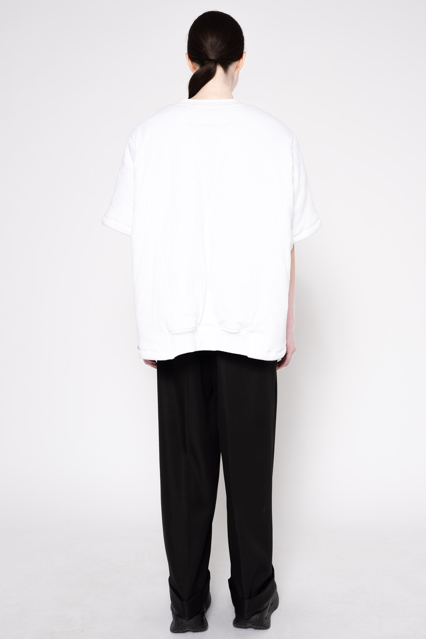 A person shown from the back wearing an oversized white short-sleeve T-shirt and loose black pants against a plain light gray studio background.