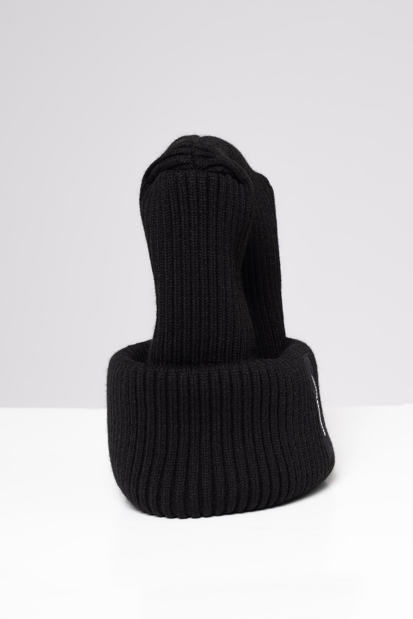 A black ribbed knit beanie hat with a folded cuff, featuring a small black vertical label on the side with white text that reads 'MELITTA BAUMEISTER'. The hat is photographed on a clean, light gray studio background.