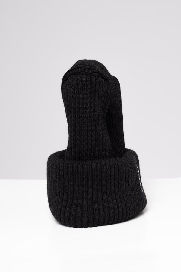 A black ribbed knit beanie hat with a folded cuff, featuring a small black vertical label on the side with white text that reads 'MELITTA BAUMEISTER'. The hat is photographed on a clean, light gray studio background.