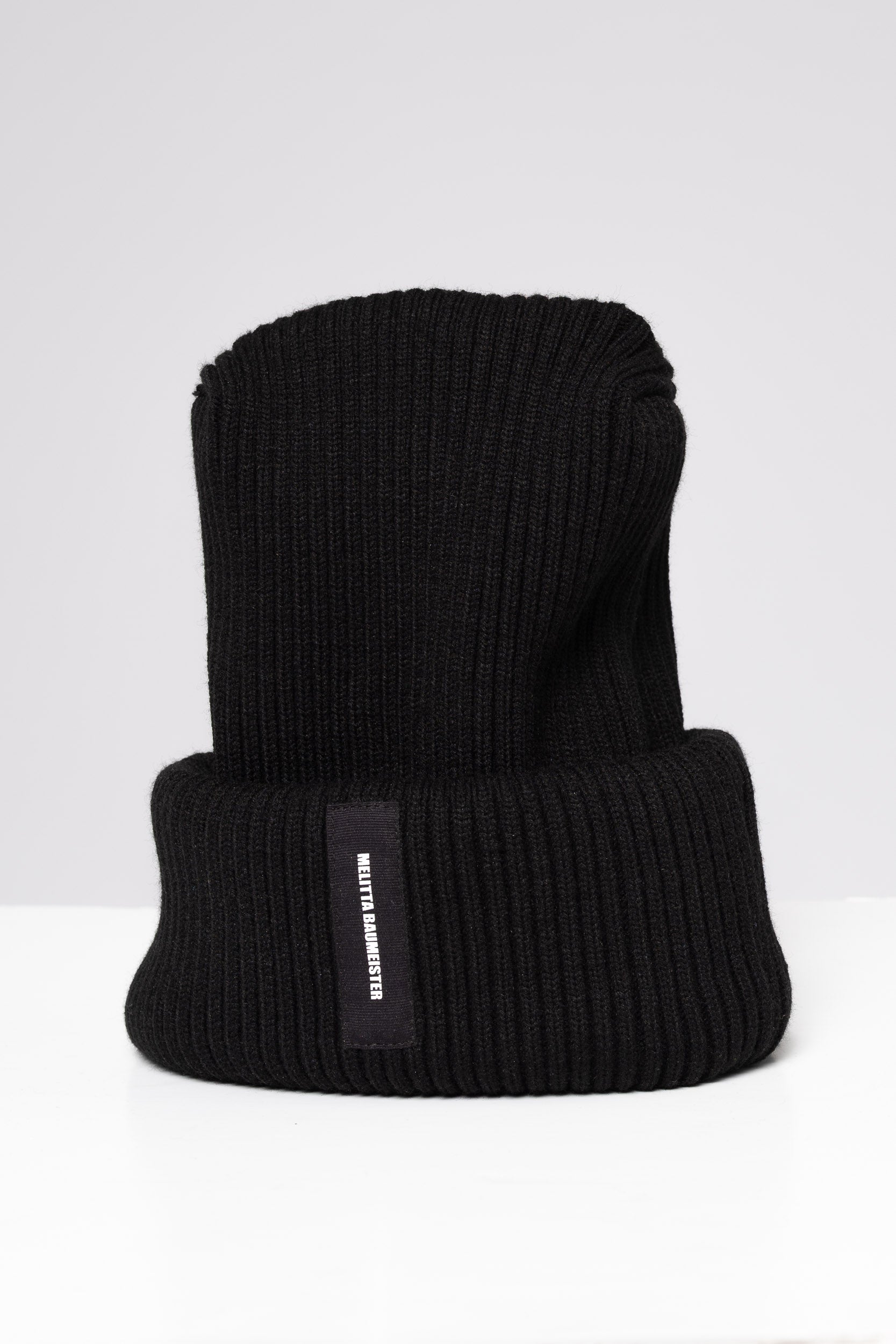 A black ribbed knit beanie hat with a folded cuff, featuring a small black vertical label on the side with white text that reads 'MELITTA BAUMEISTER'. The hat is photographed on a clean, light gray studio background.