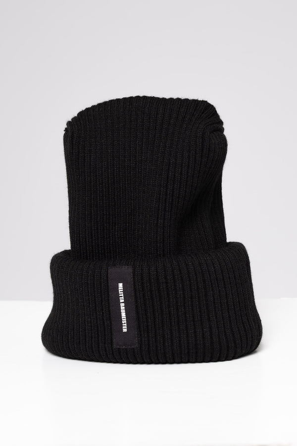A black ribbed knit beanie hat with a folded cuff, featuring a small black vertical label on the side with white text that reads 'MELITTA BAUMEISTER'. The hat is photographed on a clean, light gray studio background.