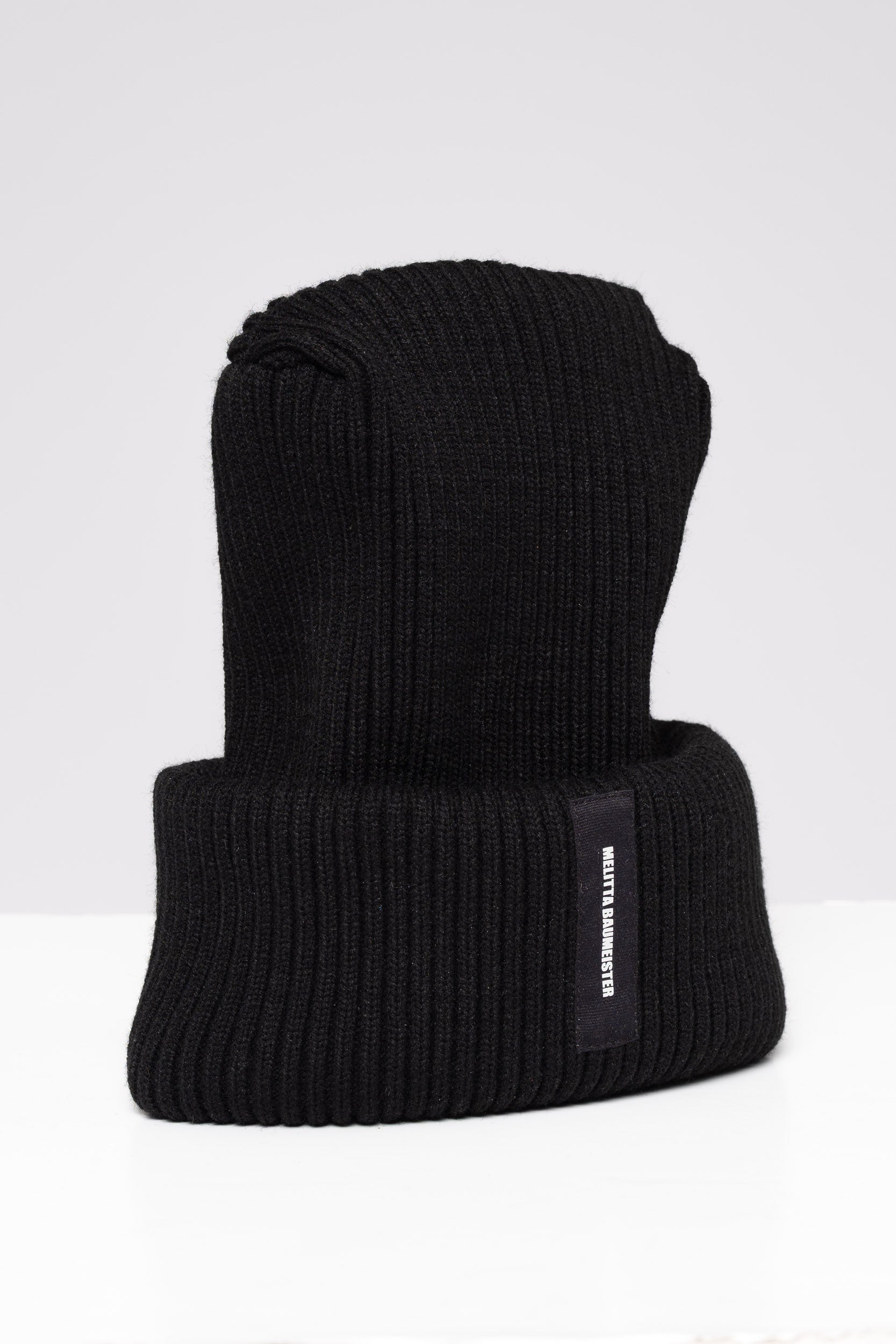 A black ribbed knit beanie hat with a folded cuff, featuring a small black vertical label on the side with white text that reads 'MELITTA BAUMEISTER'. The hat is photographed on a clean, light gray studio background.