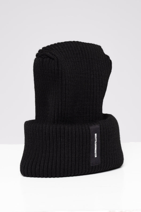 A black ribbed knit beanie hat with a folded cuff, featuring a small black vertical label on the side with white text that reads 'MELITTA BAUMEISTER'. The hat is photographed on a clean, light gray studio background.