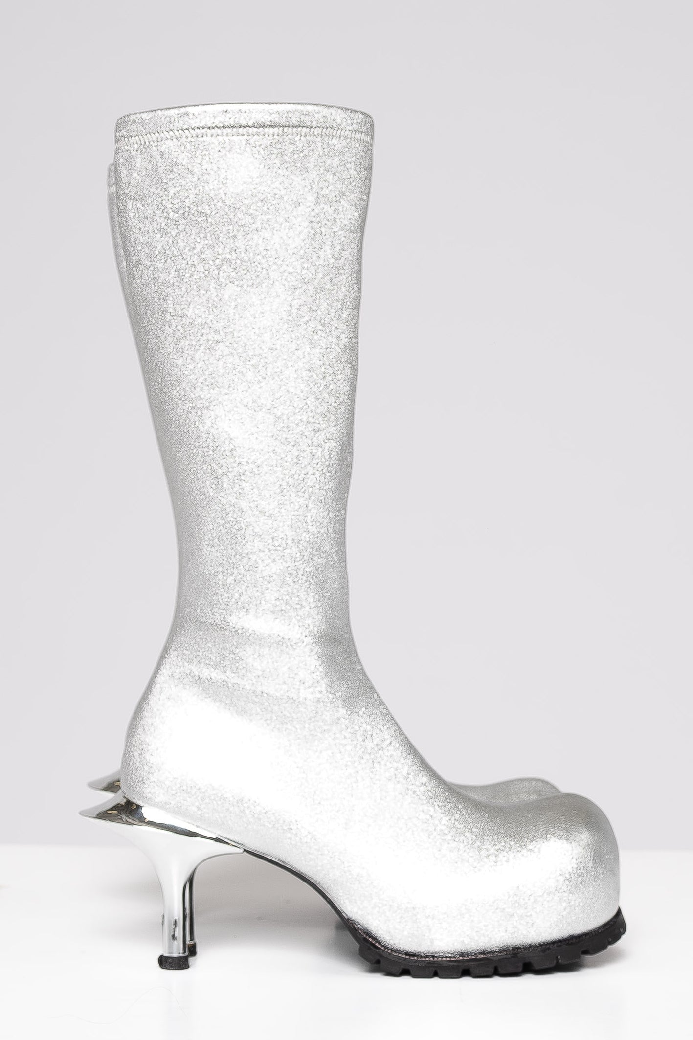 A single knee-high boot covered in sparkling silver glitter, featuring a chunky silver high heel with a metallic spike detail and a black treaded sole, shown against a light gray background.