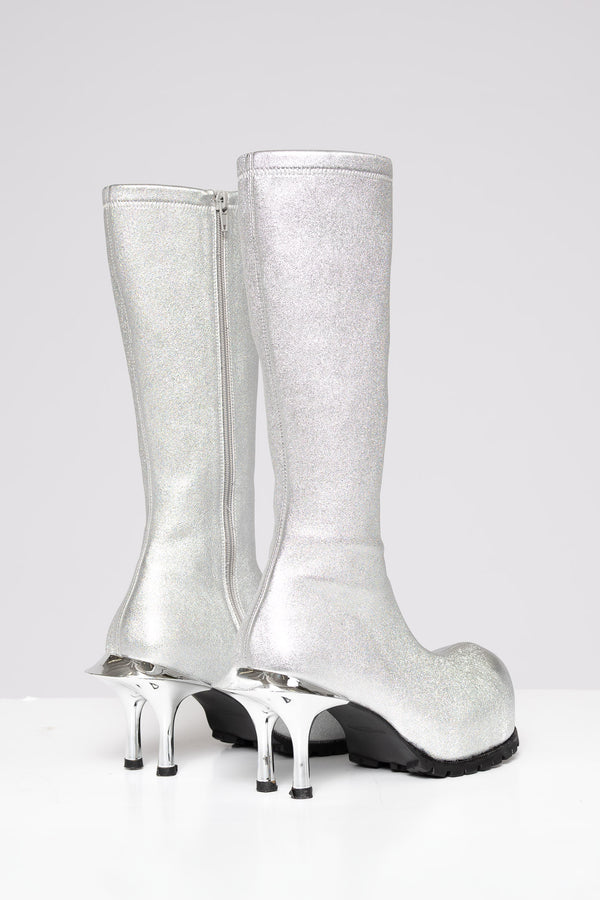 A pair of knee-high silver glitter boots with unique metallic silver high heels and black soles, shown against a plain light gray background.