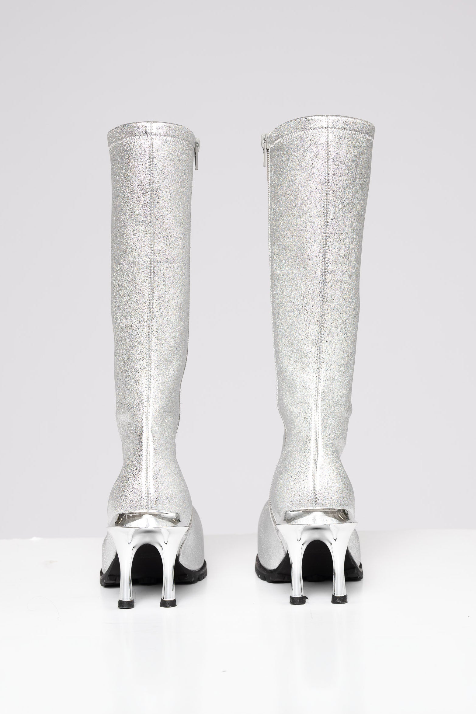 A pair of metallic silver knee-high boots with unique high heels, shown from the back on a light gray background.
