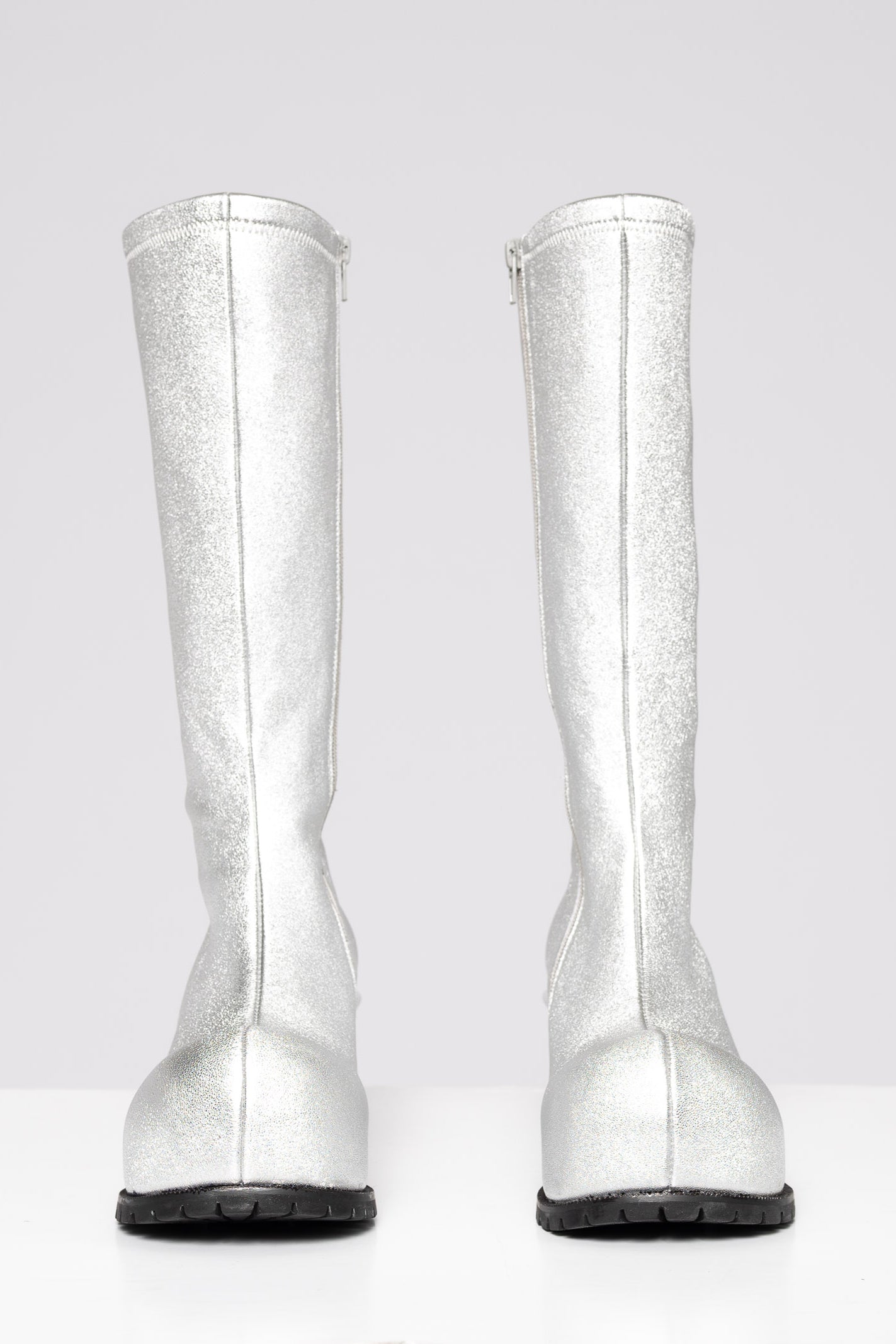 A pair of metallic silver knee-high boots shown from the back, featuring a textured glitter finish and black soles, set against a plain light gray background.