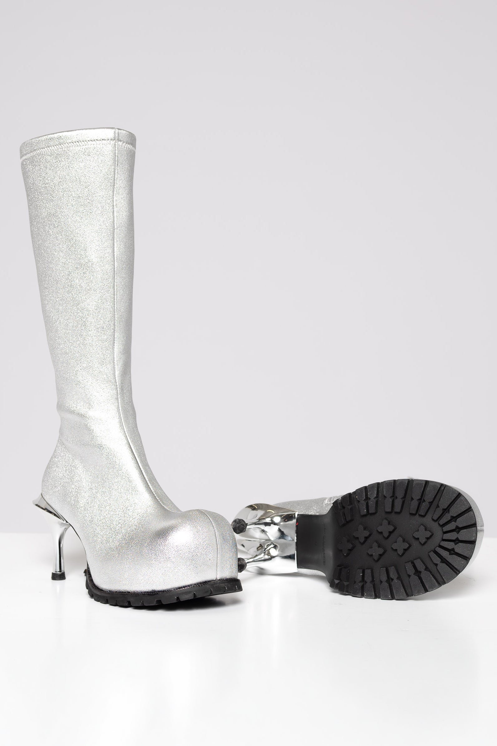 A metallic silver knee-high boot with a double high stiletto heel, shown alongside its detached black treaded sole on a light gray background.