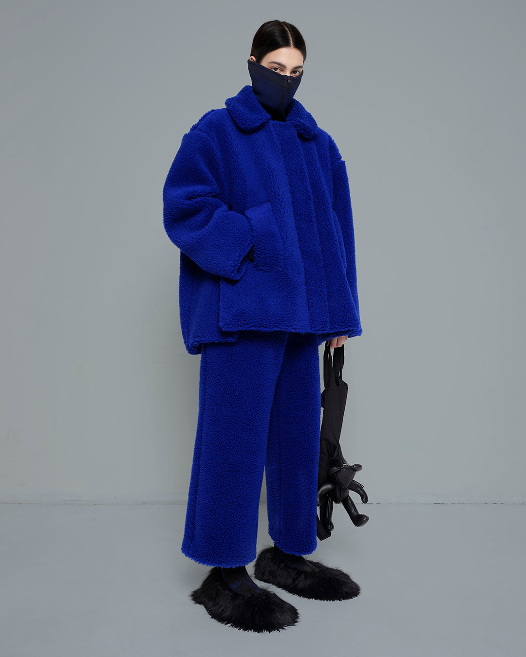 MELITTA-BAUMEISTER-BLACK-TEDDY-TOTE-BAG-LONG-BLACK-HAIR-FEMALE-MODEL-HOLDING-THE-BAG-WHILE-WEARING-BLUE-SOFT-LOOKING-JACKET-AND-PANTS-WITH-BLACK-FURRY-SHOES
