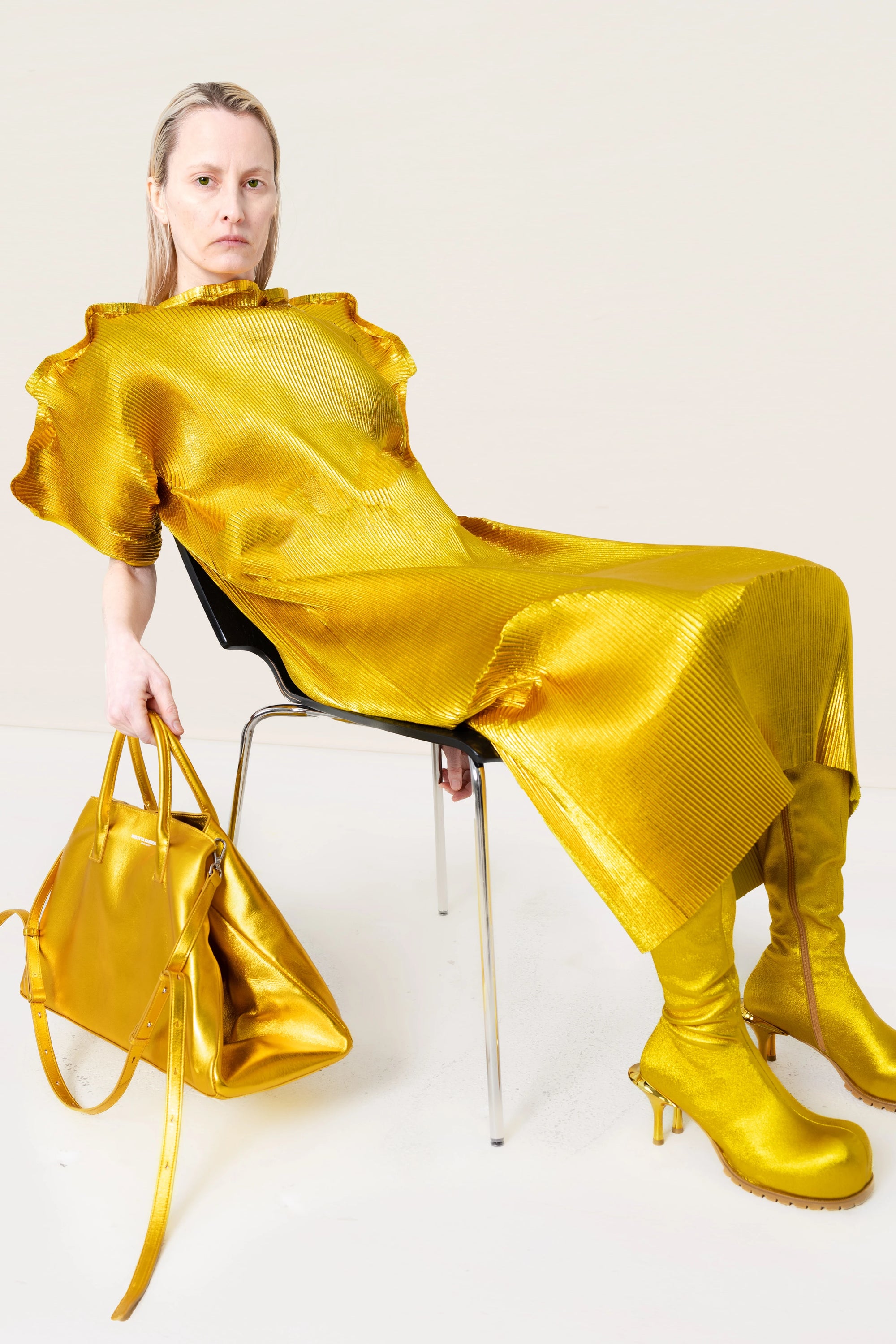 PERSON-SITTING-BACK-IN-CHAIR-WEAR-GOLD-RIPPLE-PLEAT-DRESS-AND-GOLD-COWHIDE-DOUBLE-HEELED-BOOTS-HOLDING-GOLD-A-LINE-SHOPPER-BYSHOULDER-STRAPS
