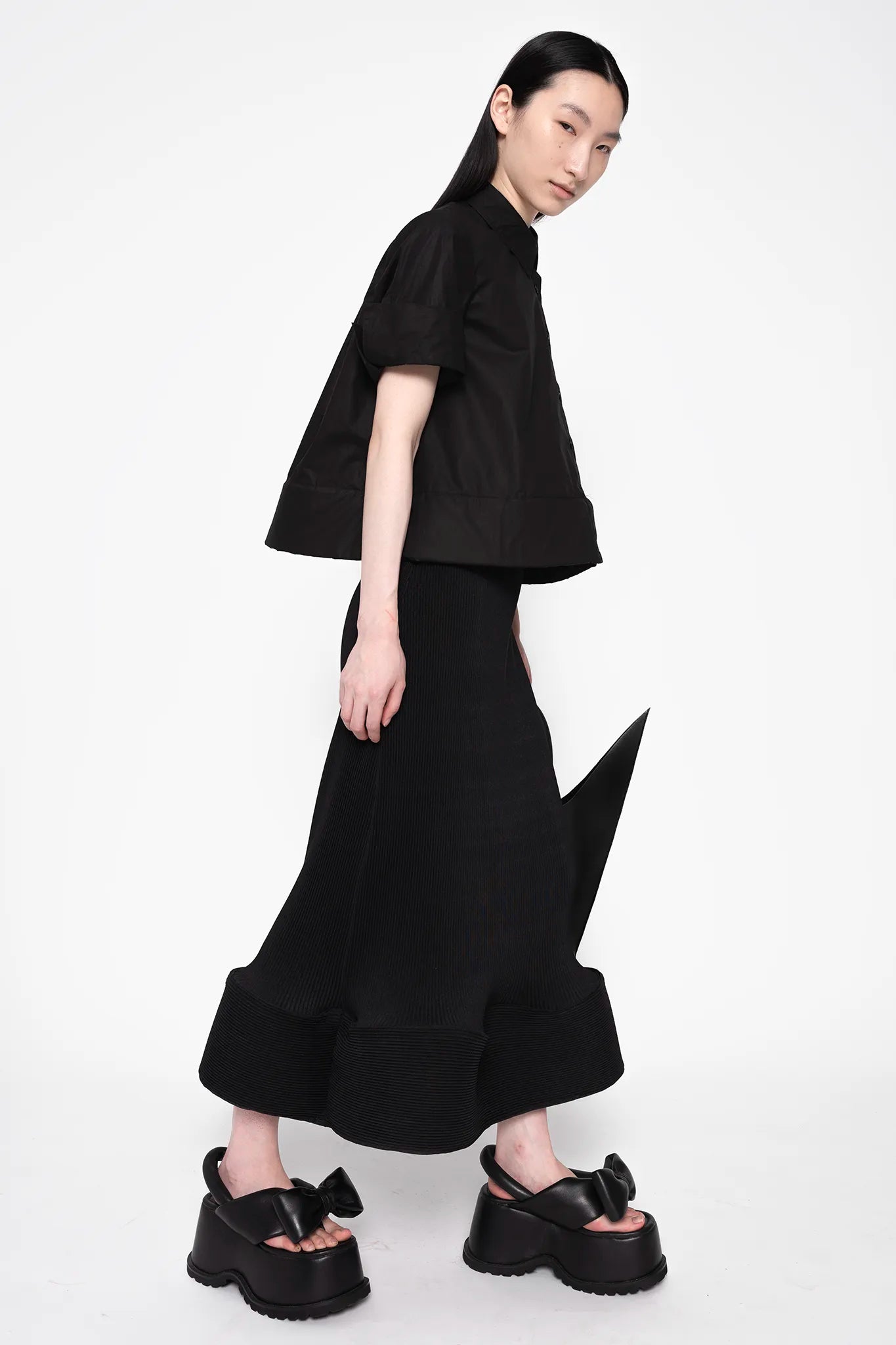 MELITTA-BAUMEISTER-Ripple-Wavy-Skirt-Black-LONG-BLACK-HAIR-FEMALE-MODEL-ALSO-WEARING-BLACK-TOP-AND-BOW-SHOES-WITH-POINTY-SHOULDER-BAG-IN-LEFT-HAND