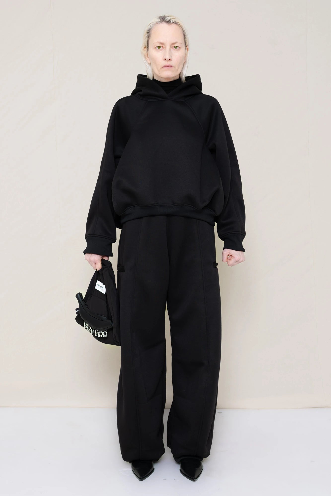 PERSON-WEARING-BLACK-CROPPED-HOODIE-WITH-MATCHING-BLACK-ROUND-LEG-LOUNGEPANTS-AND-BLACK-FLAT-POINTY-BOOTS-HOLDING-PIERCED-BLACK-BANANA-TOTE-BAG-FRONT-VIEW