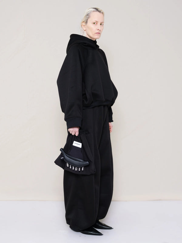 PERSON-WEARING-BLACK-CROPPED-HOODIE-WITH-MATCHING-BLACK-ROUND-LEG-LOUNGEPANTS-AND-BLACK-FLAT-POINTY-BOOTS-HOLDING-PIERCED-BLACK-BANANA-TOTE-BAG-THREE-QUARTER-VIEW