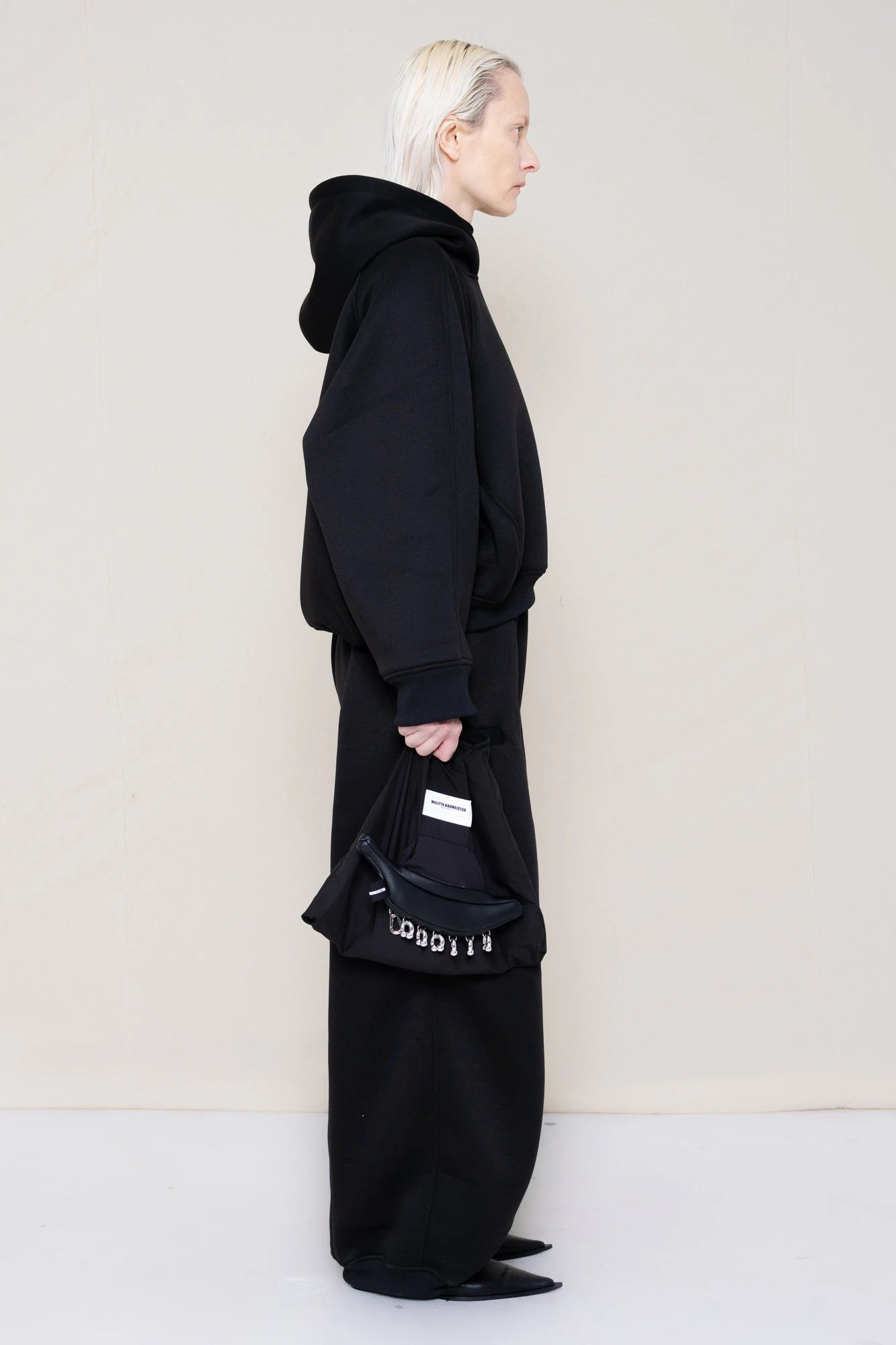 PERSON-WEARING-BLACK-CROPPED-HOODIE-WITH-MATCHING-BLACK-ROUND-LEG-LOUNGEPANTS-AND-BLACK-FLAT-POINTY-BOOTS-HOLDING-PIERCED-BLACK-BANANA-TOTE-BAG-SIDE-VIEW