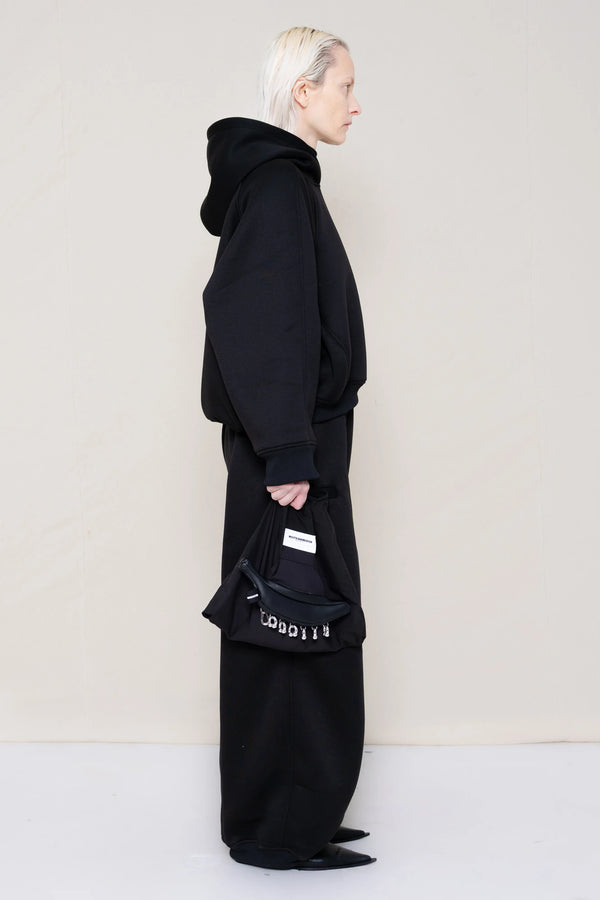 PERSON-WEARING-BLACK-CROPPED-HOODIE-WITH-MATCHING-BLACK-ROUND-LEG-LOUNGEPANTS-AND-BLACK-FLAT-POINTY-BOOTS-HOLDING-PIERCED-BLACK-BANANA-TOTE-BAG-SIDE-VIEW
