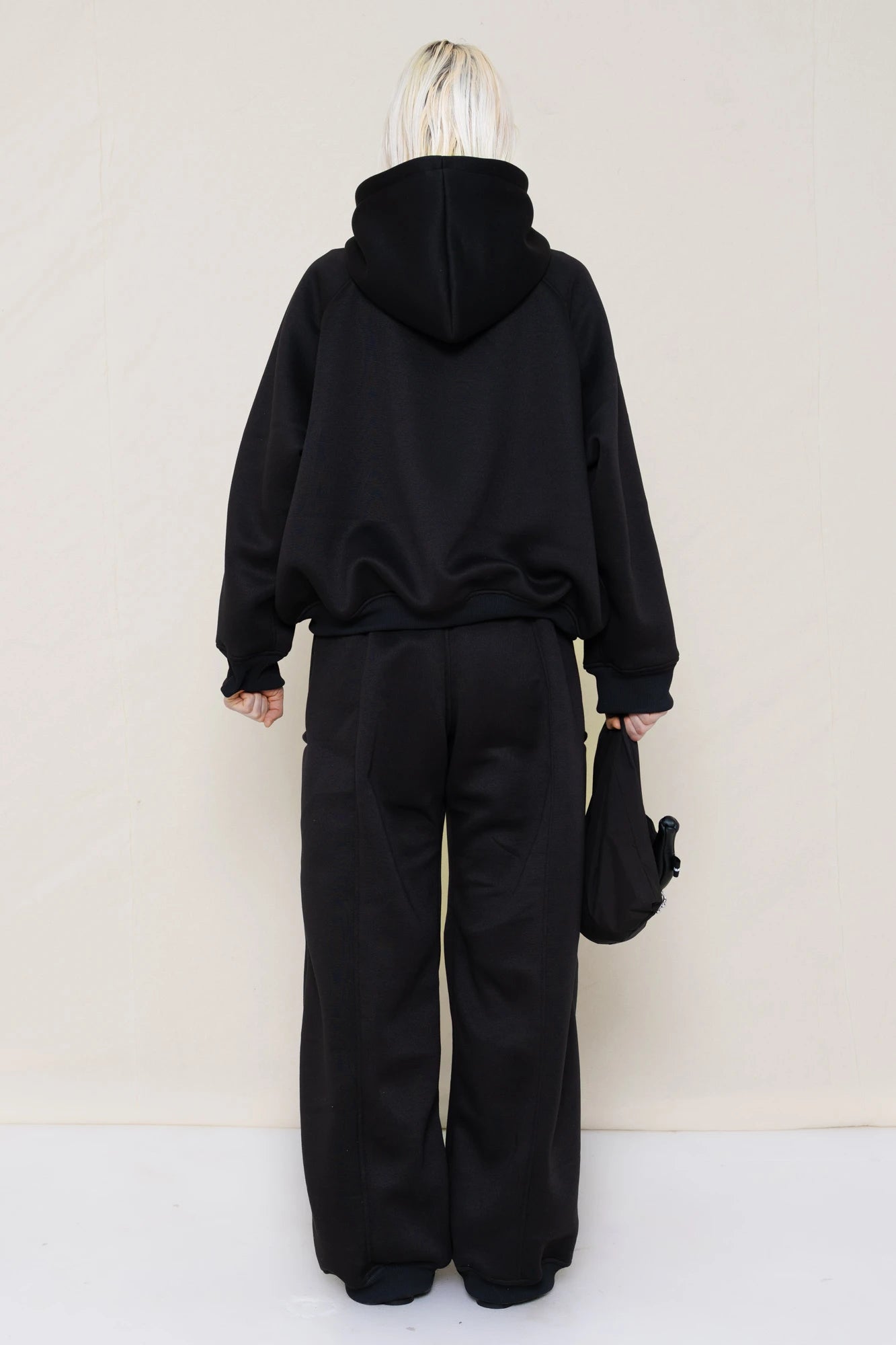 PERSON-WEARING-BLACK-CROPPED-HOODIE-WITH-MATCHING-BLACK-ROUND-LEG-LOUNGEPANTS-AND-BLACK-FLAT-POINTY-BOOTS-HOLDING-PIERCED-BLACK-BANANA-TOTE-BAG-BACK-VIEW