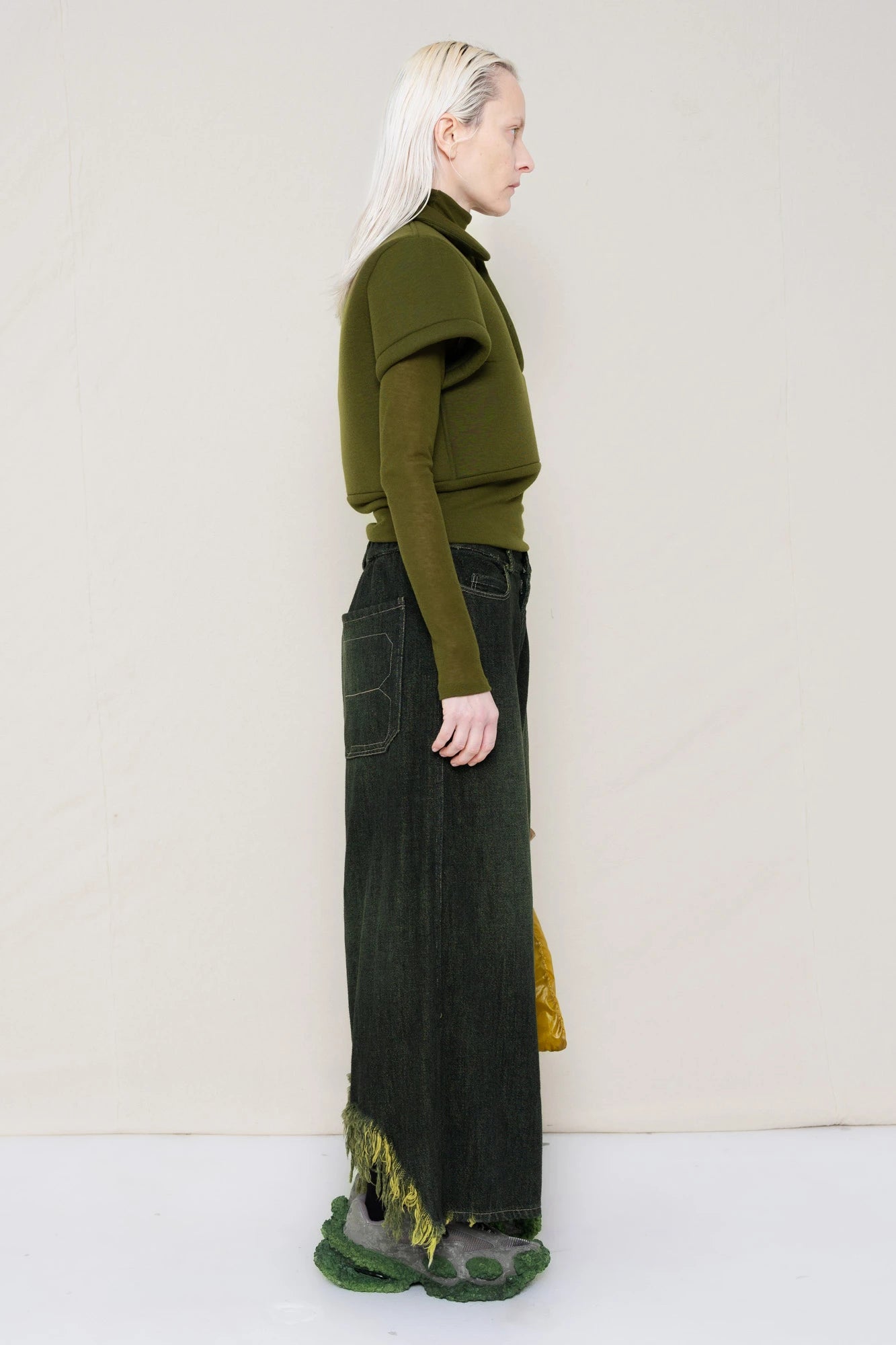 PERSON-WEARING-MOSS-FOAMY-POLO-WITH-MOSS-LONG-SLEEVE-TURTLENECK-UNDER-AND-BLACK-YELLOW-WASH-DENIM-PANTS-WITH-CUSTOMIZED-SNEAKERS-HOLDING-GOLD-BANANA-TOTE-BAG-SIDE-VIEW