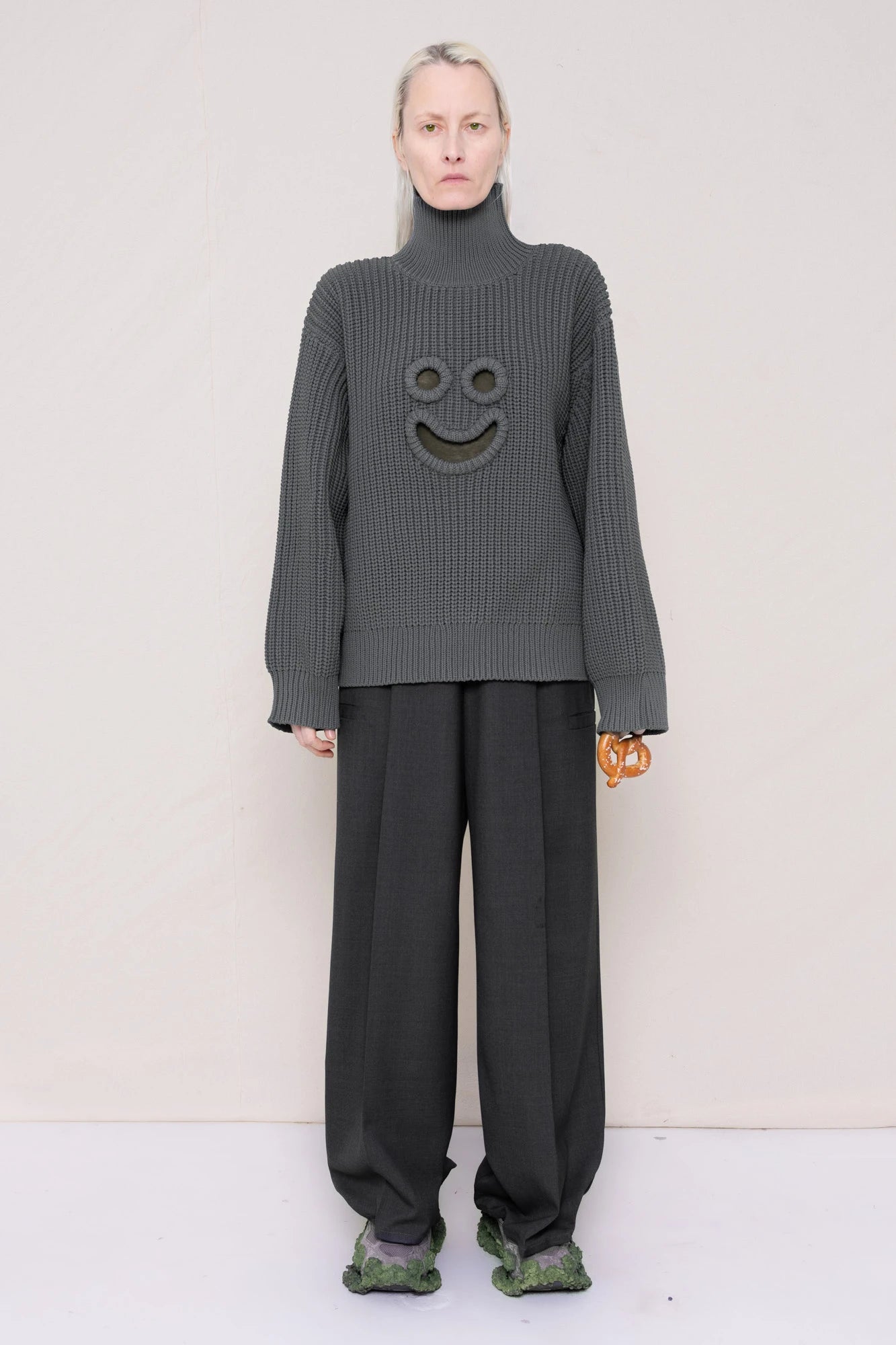 woman-holding-a-pretzel-wearing-a-gray-knit-turtle-neck-sweater-with-a-smiley-face-cut-out-and-grey-WOOL-BLEND-TWILL-TAILORED-PANTS-WITH-ZIGZAG-HEM-DETAIL-worn-with-custom-shoes-full-body-front-view
