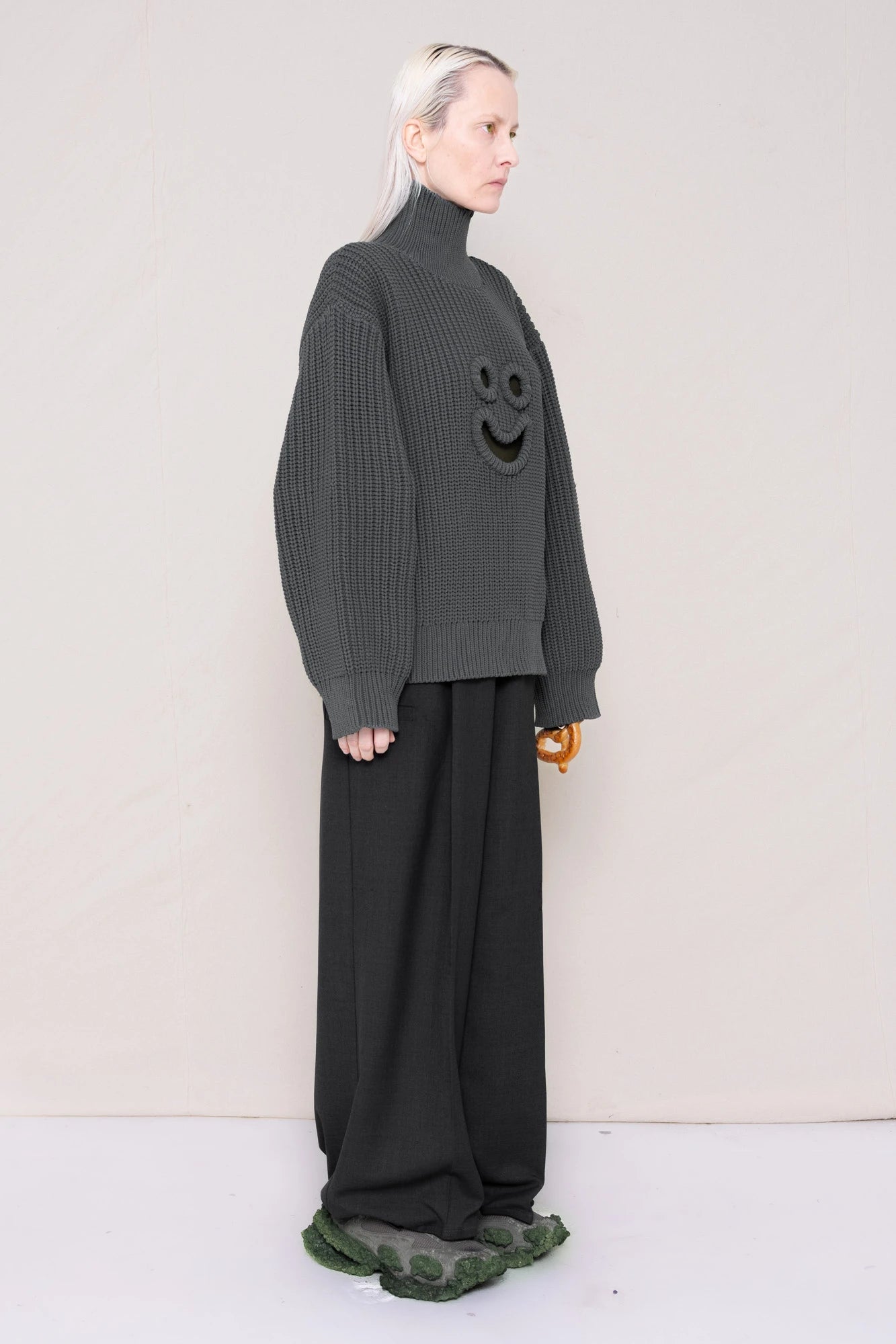 woman-holding-a-pretzel-wearing-a-gray-knit-turtle-neck-sweater-with-a-smiley-face-cut-out-and-grey-WOOL-BLEND-TWILL-TAILORED-PANTS-WITH-ZIGZAG-HEM-DETAIL-worn-with-custom-shoes-full-body-three-quarters-view