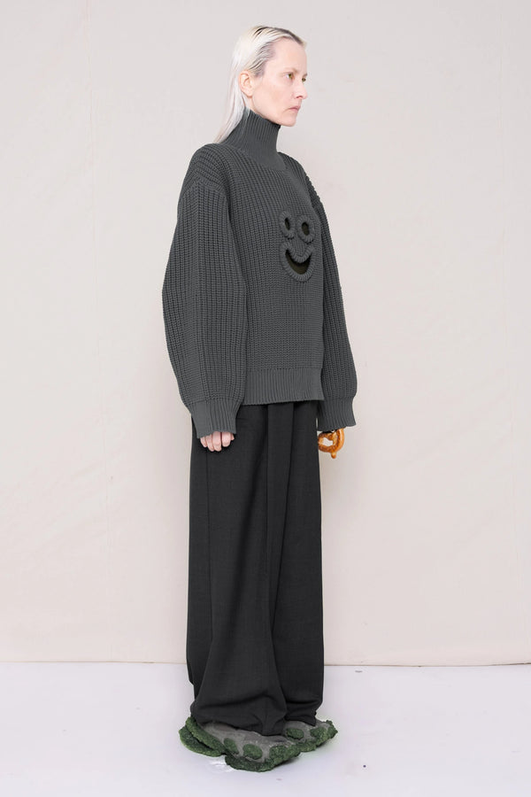 woman-holding-a-pretzel-wearing-a-gray-knit-turtle-neck-sweater-with-a-smiley-face-cut-out-and-grey-WOOL-BLEND-TWILL-TAILORED-PANTS-WITH-ZIGZAG-HEM-DETAIL-worn-with-custom-shoes-full-body-three-quarters-view