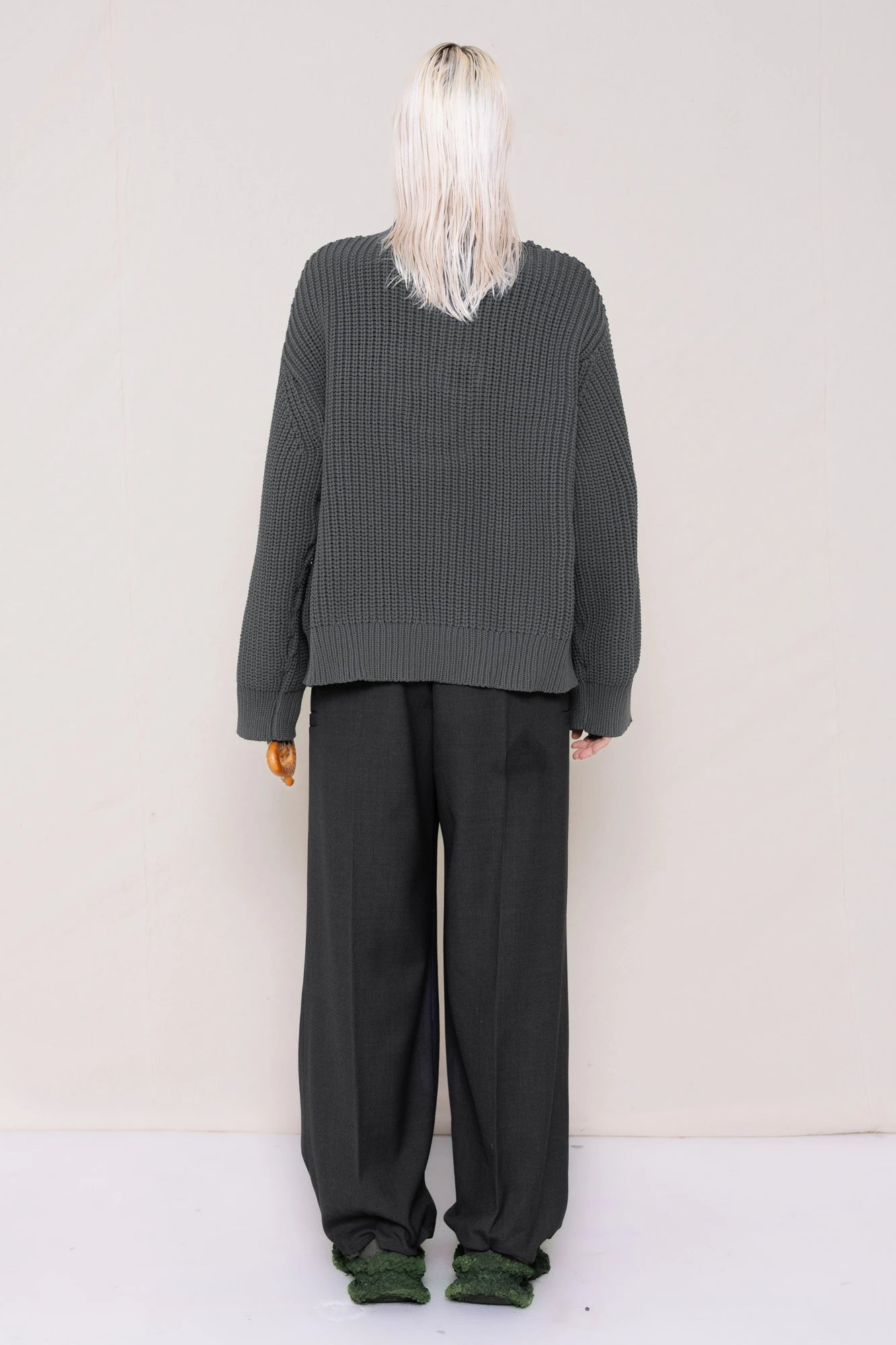 woman-holding-a-pretzel-wearing-a-gray-knit-turtle-neck-sweater-with-a-smiley-face-cut-out-and-grey-WOOL-BLEND-TWILL-TAILORED-PANTS-WITH-ZIGZAG-HEM-DETAIL-worn-with-custom-shoes-full-body-back-view