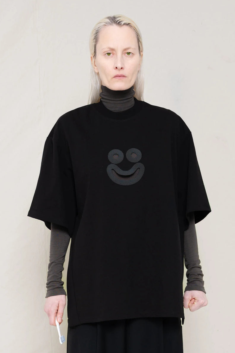 PERSON-WITH-GREEN-EYES-WEARING-BLACK-OVERSIZE-HAPPY-SMILEY-SHIRT-OVER-GREY-TURTLENECK-LONGSLEEVE-AND-WEARS-BLACK-WIDE-TROUSERS-AND-MUD-CUSTOMIZED-NIKE-SNEAKER-IN-GREY-HOLDING-TOOTHBRUSH-FRONT-VIEW